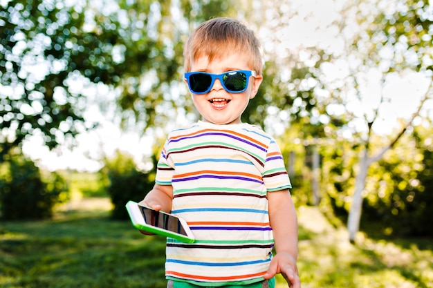 Маленький милый мальчик держит телефон в руке и улыбается в летнем саду.