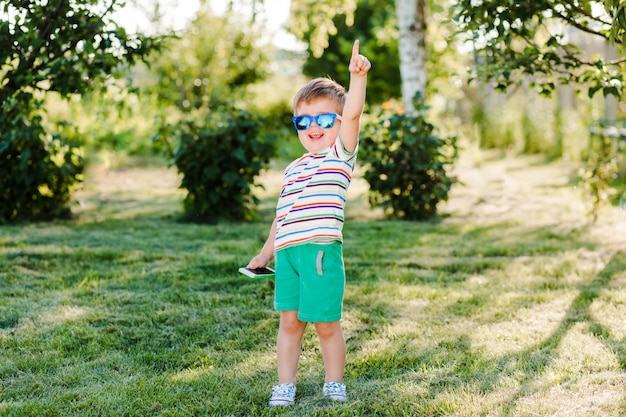 Маленький милый мальчик выглядит вдохновленным и счастливым в ярких солнцезащитных очках со своим телефоном.