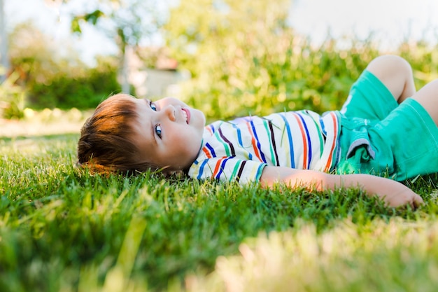 Little cute boy resting on the green grass in the garden and looks happy and relaxed.