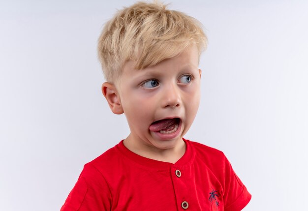 Маленький милый мальчик со светлыми волосами и голубыми глазами в красной футболке смотрит сбоку, показывая язык на белой стене
