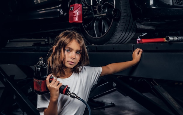 小さなかわいい子供は、車のワークショップで空気圧ドリルを渡す写真家のためにポーズをとっています。