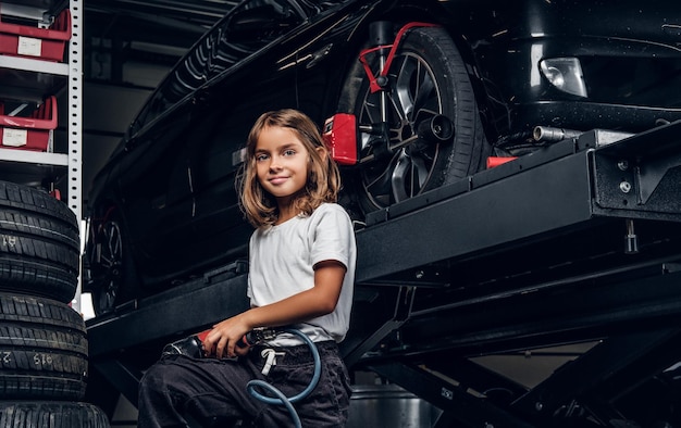 작은 귀여운 아이가 자동차 작업장에서 공압 드릴을 나눠주는 사진사를 위해 포즈를 취하고 있습니다.
