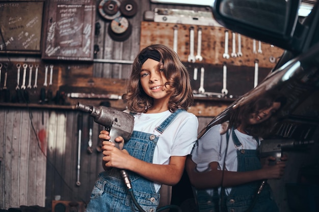 La piccola ragazza sveglia sta posando con il trapano pneumatico per il fotografo nell'officina del servizio automatico.