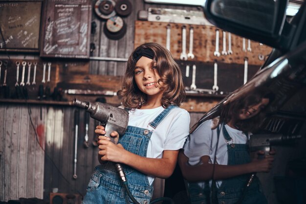 Маленькая симпатичная девочка позирует фотографу с пневматической дрелью в автосервисе.