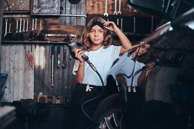 Little cute girl is posing with pneumatic drill for photographer at auto service workshop.