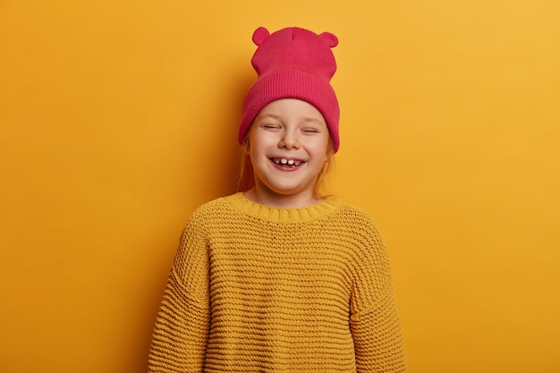 Little cute girl laughs happily, feels glad to receive new toy doll from parents, closes eyes, has fun indoor, wears hat with ears and loose knitted sweater, isolated over yellow wall.