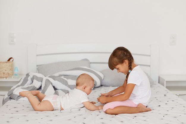 Little cute girl wearing white t shirt and pink shorts playing with her little infant sister while being together in light room, happy childhood, sibling spending time together.