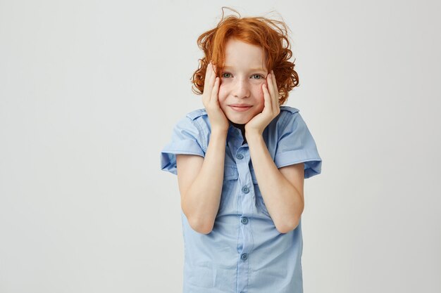 Бесплатное фото Маленький милый ребенок с рыжими вьющимися волосами и веснушками, держа голову руками и улыбается