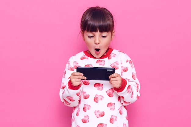 Free photo little dark haired charming girl with dark hair holding smart phone with opened mouth, playing games, being surprised of result, posing isolated over pink wall.