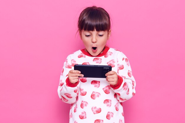 Little dark haired charming girl with dark hair holding smart phone with opened mouth, playing games, being surprised of result, posing isolated over pink wall.