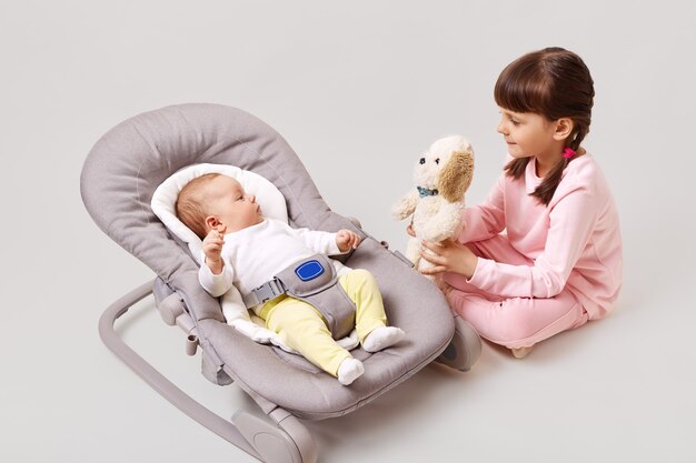 A little dark-haired girl with pigtails is playing with her newborn sister or brother who is lying in a baby rocking chair