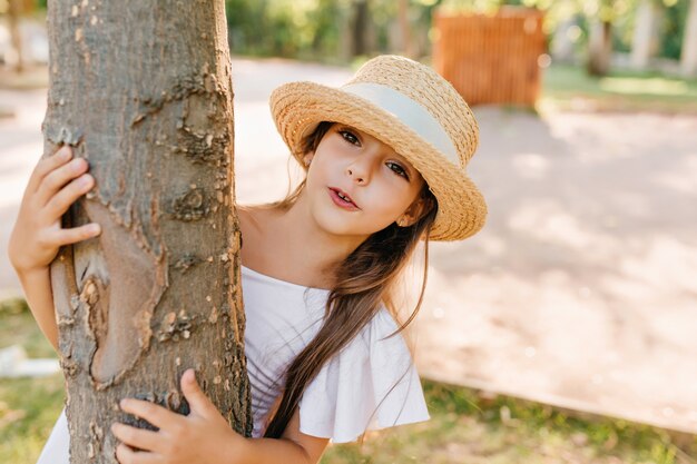 Little fashionable lady hiding behind tree, while playing in park in summer day. Pretty brunette girl in hat with white ribbon and elegant dress spending vacation outdoor.