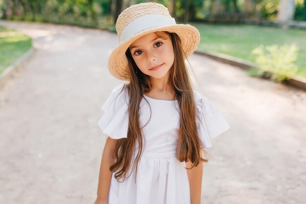 Маленькая модная дама с длинными ресницами с интересом смотрит, стоя на дороге в элегантной шляпе. Открытый портрет застенчивой шатенки в милом белом платье.