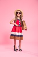 little fashionista with a shopping bag in a summer hat and glasses , on a colored pink background in mom's shoes , the concept of children's fashion