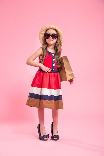 子供のファッションのコンセプト、ママの靴のピンク色の背景に夏の帽子とメガネのショッピングバッグを持つ小さなファッショニスタ