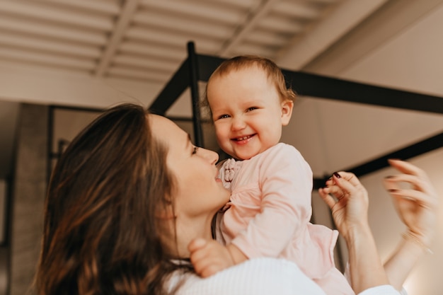 Little female baby laughs and looks into camera while mom hugs her and holds her in arms.