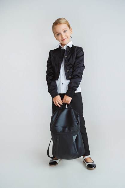 Little female model posing in school's uniform with backpack on white wall