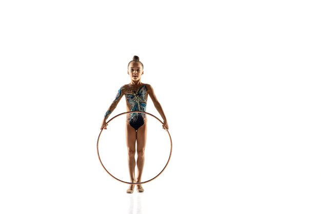 Маленькая гибкая девочка изолированная на белой стене. Маленькая женская модель в роли артистки художественной гимнастики в ярком купальнике. Изящество в движении, действии и спорте. Выполнение упражнений с обручем.