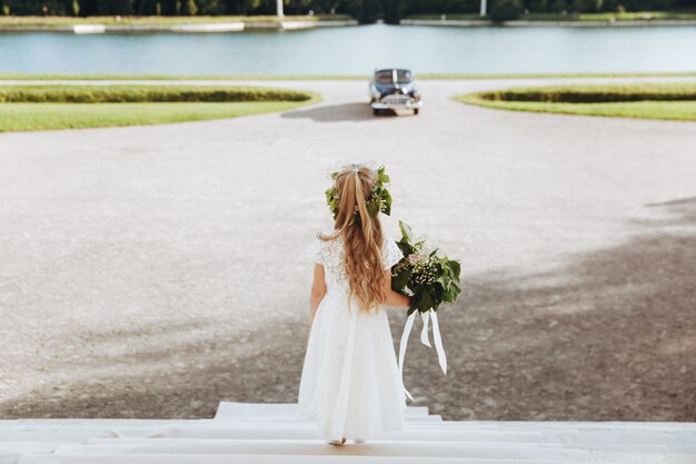 Little flower girl looks at black retro car riding towards the house