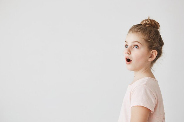 Little funny girl with blonde hair in bun standing with open mouth on street, being shocked seeing fire in neighbor's home.