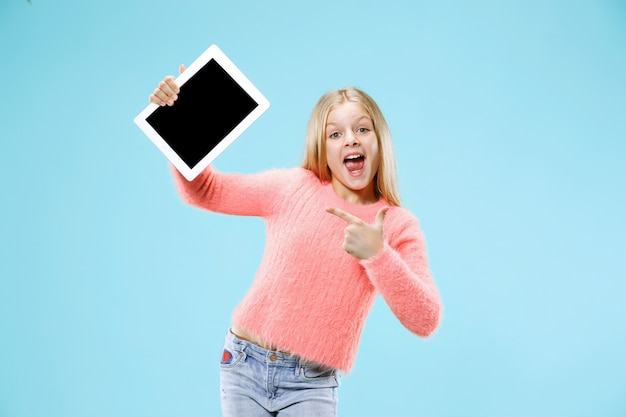 Little funny girl with tablet on blue studio background. She showing something and pointing at screen.