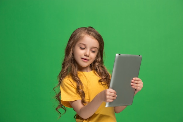 緑のスタジオの背景にタブレットを持つ小さな面白い女の子。彼女は何かを見せて画面を見ています。