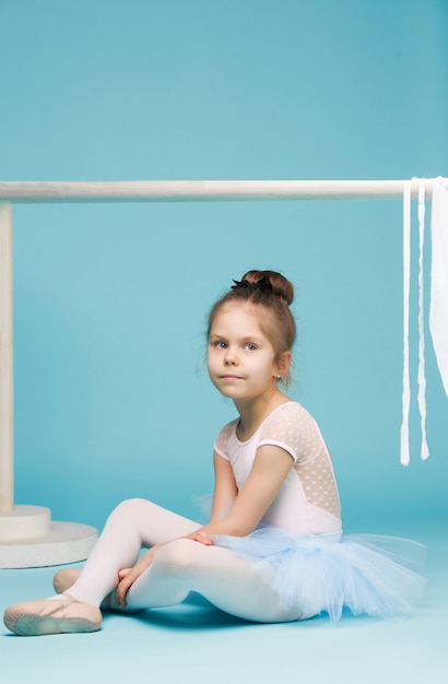 The little girl as balerina dancer sitting and posing near ballet rack on blue studio