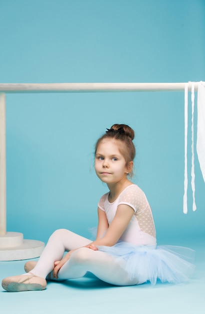 Маленькая девочка как балерина сидит и позирует возле балетной стойки на синей студии