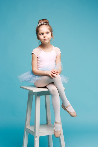 La bambina come ballerina della ballerina che si siede sulla sedia di legno bianca allo studio blu