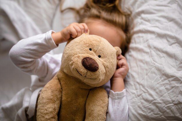 柔らかいおもちゃが付いているベッドの小さな女の子、白いベッドの子供の感情