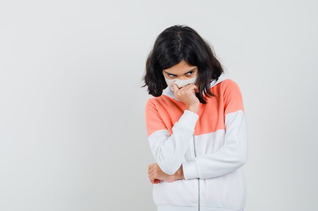 Little girl bending head down, holding hand on mouth in jacket, mask and looking upset. front view.