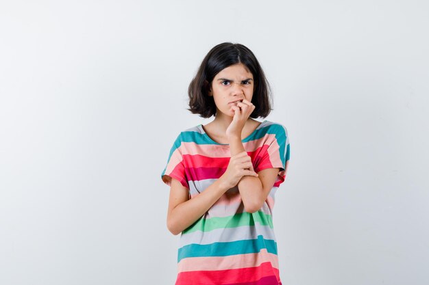 Little girl biting fingers while holding hand on forearm in t-shirt, jeans and looking agitated , front view.