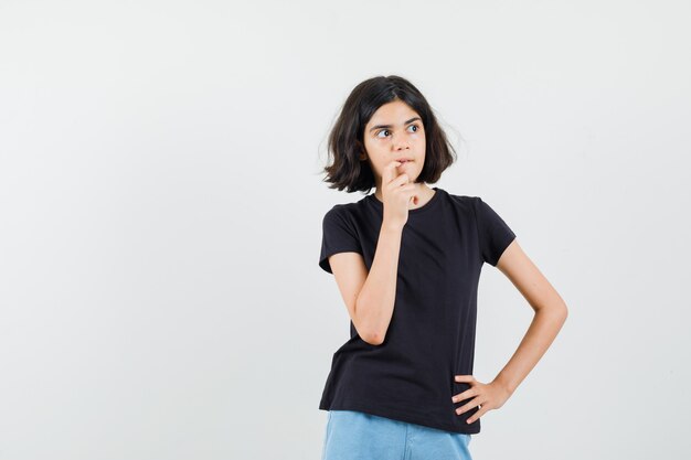 Little girl biting nail in black t-shirt, shorts and looking thoughtful. front view.