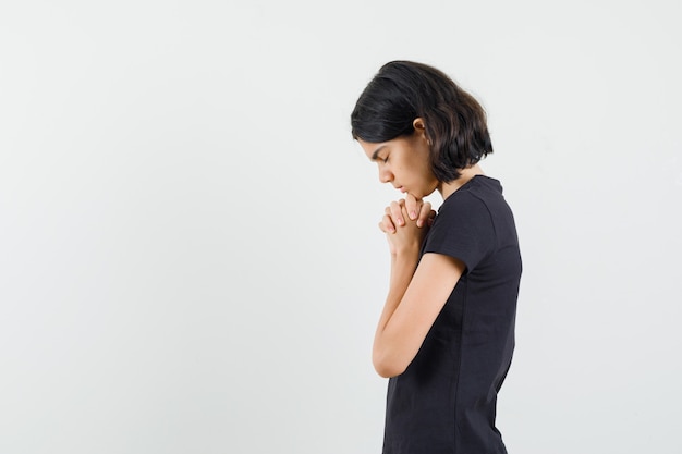 Little girl in black t-shirt clasping hands in praying gesture and looking hopeful .