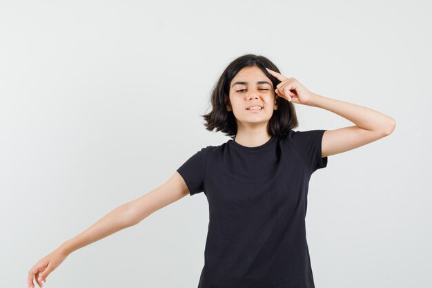 Little girl in black t-shirt holding finger on head, stretching arm and looking pensive , front view.