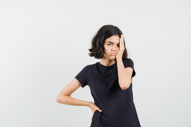 Little girl in black t-shirt holding hand face and looking confused , front view.