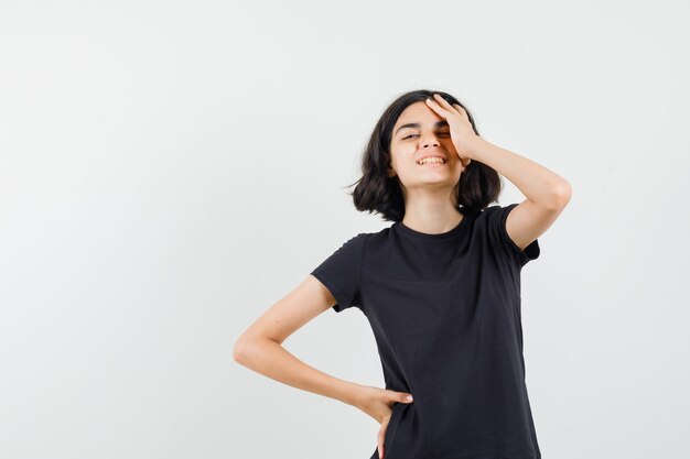 Little girl in black t-shirt holding hand on forehead and looking pretty , front view.