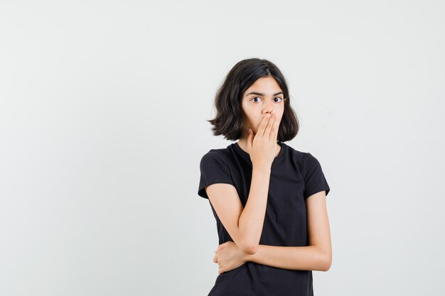 Little girl in black t-shirt holding hand on mouth and looking wondered , front view.