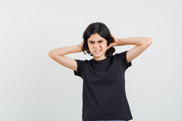 Little girl in black t-shirt holding hands on hair and looking discontent , front view.