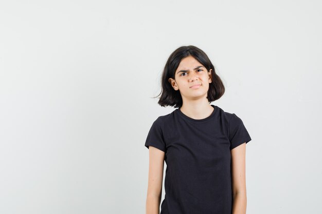 Little girl in black t-shirt looking at front and looking discontent , front view.