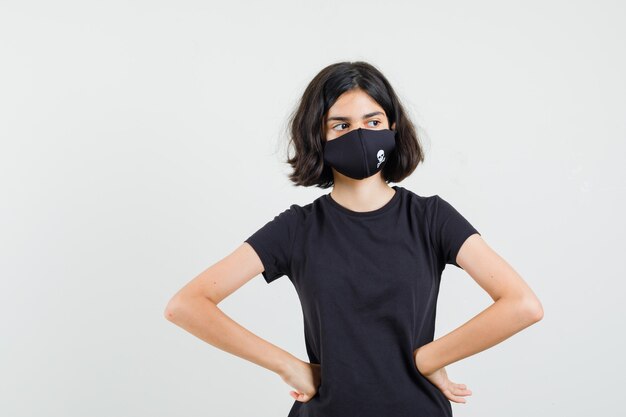 Little girl in black t-shirt, mask holding hands on waist and looking serious , front view.