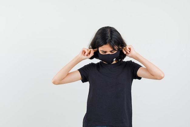 Little girl in black t-shirt, mask plugging ears with fingers and looking annoyed , front view.