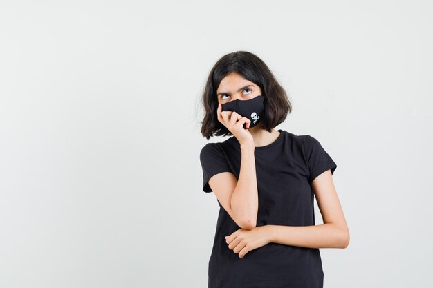 Маленькая девочка в черной футболке, маска стоя в позе мышления, вид спереди.
