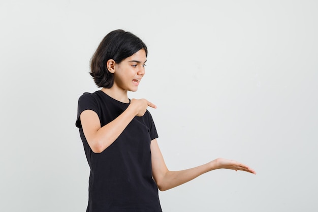 Little girl in black t-shirt pointing at her palm spread and looking cheery , front view.