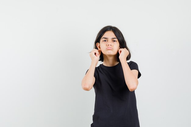 Little girl in black t-shirt pulling down her earlobes and looking offended , front view.