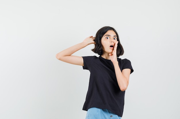 Little girl in black t-shirt, shorts holding hand near open mouth and looking surprised , front view.