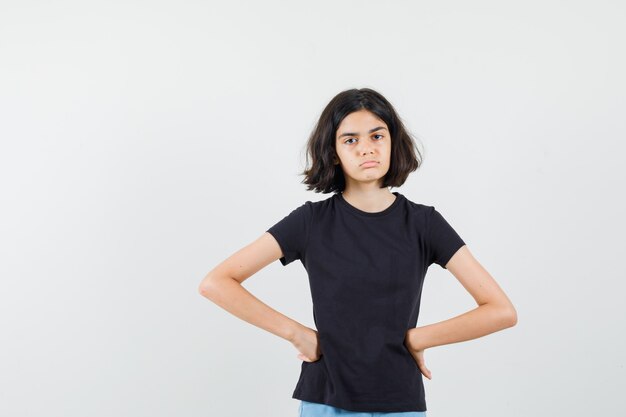 Маленькая девочка в черной футболке, шортах, взявшись за руки на талии и грустно, вид спереди.