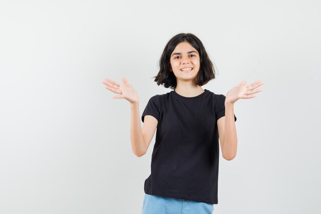 Little girl in black t-shirt, shorts raising open palms and looking cheerful , front view.