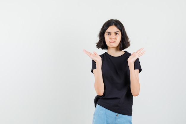 Little girl in black t-shirt, shorts raising open palms and looking gloomy , front view.