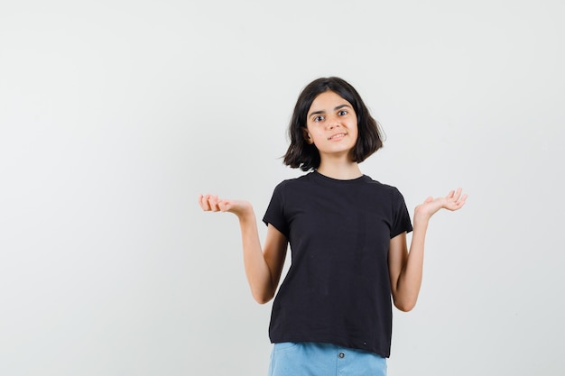 Little girl in black t-shirt, shorts showing helpless gesture and looking confused , front view.